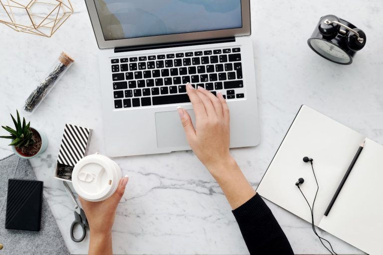 Top view of a woman using a laptop with coffee, notebook, and accessories on a desk.